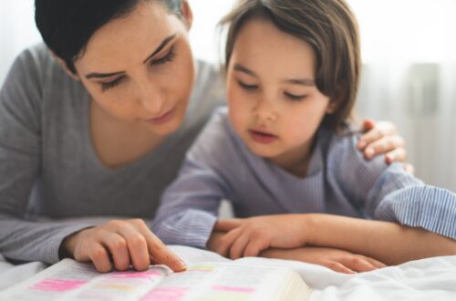 A woman and child kneeling beside a bed while reading an open book together in a bright bedroom.
