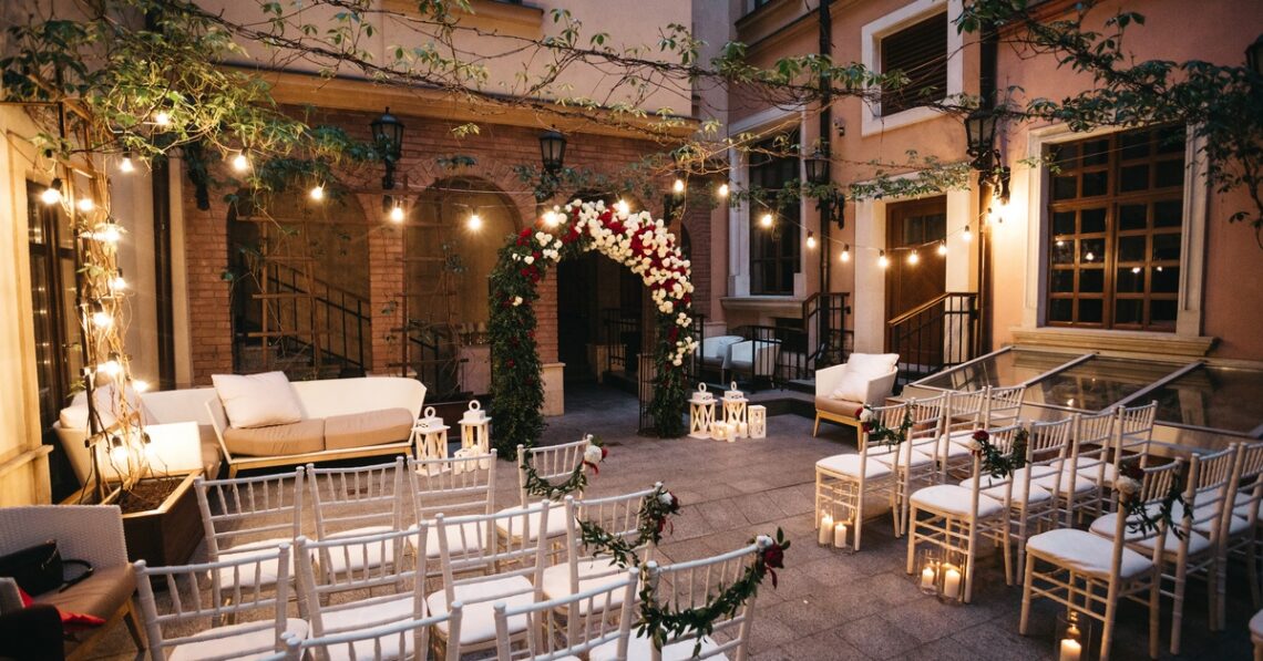A courtyard wedding aisle with white chairs, floral garlands, candle lanterns, string lights, and a flower arch.