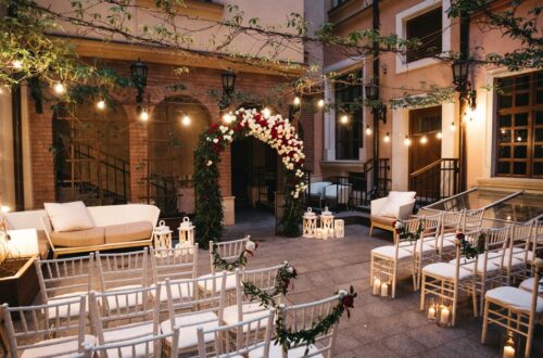 A courtyard wedding aisle with white chairs, floral garlands, candle lanterns, string lights, and a flower arch.