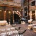 A courtyard wedding aisle with white chairs, floral garlands, candle lanterns, string lights, and a flower arch.
