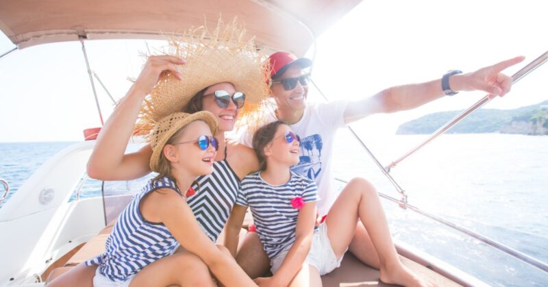 Two adults and two children wearing sunglasses and smiling on a boat as the man points toward the water.