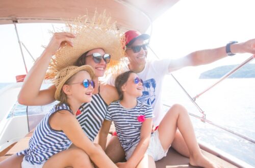 Two adults and two children wearing sunglasses and smiling on a boat as the man points toward the water.