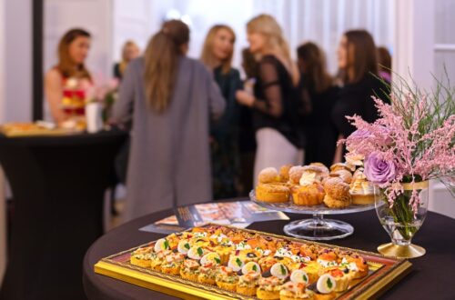 A buffet table with appetizers, pastries, and a floral arrangement in the foreground, with guests gathered behind the table.