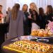 A buffet table with appetizers, pastries, and a floral arrangement in the foreground, with guests gathered behind the table.