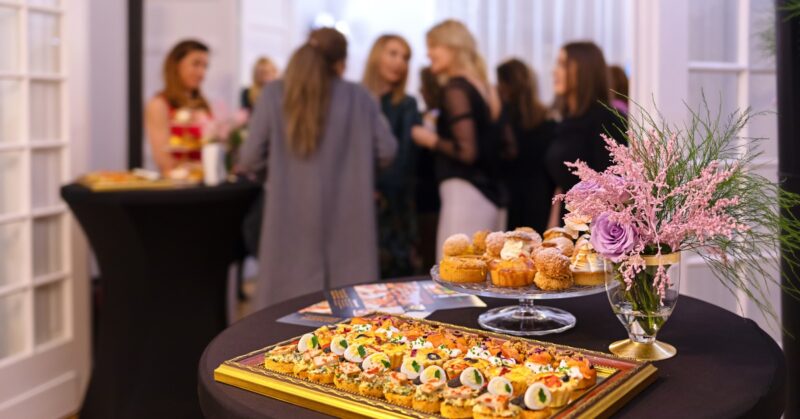A buffet table with appetizers, pastries, and a floral arrangement in the foreground, with guests gathered behind the table.