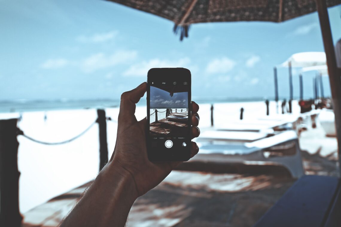 Person holding a smartphone at the beach, capturing a photo of a boardwalk and ocean under a sunny sky. Jackpot City Online Casino