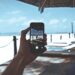 Person holding a smartphone at the beach, capturing a photo of a boardwalk and ocean under a sunny sky. Jackpot City Online Casino
