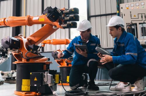 A close-up of two workers in white hard hats kneeling beside an orange robotic arm and cables near a control panel.