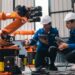 A close-up of two workers in white hard hats kneeling beside an orange robotic arm and cables near a control panel.