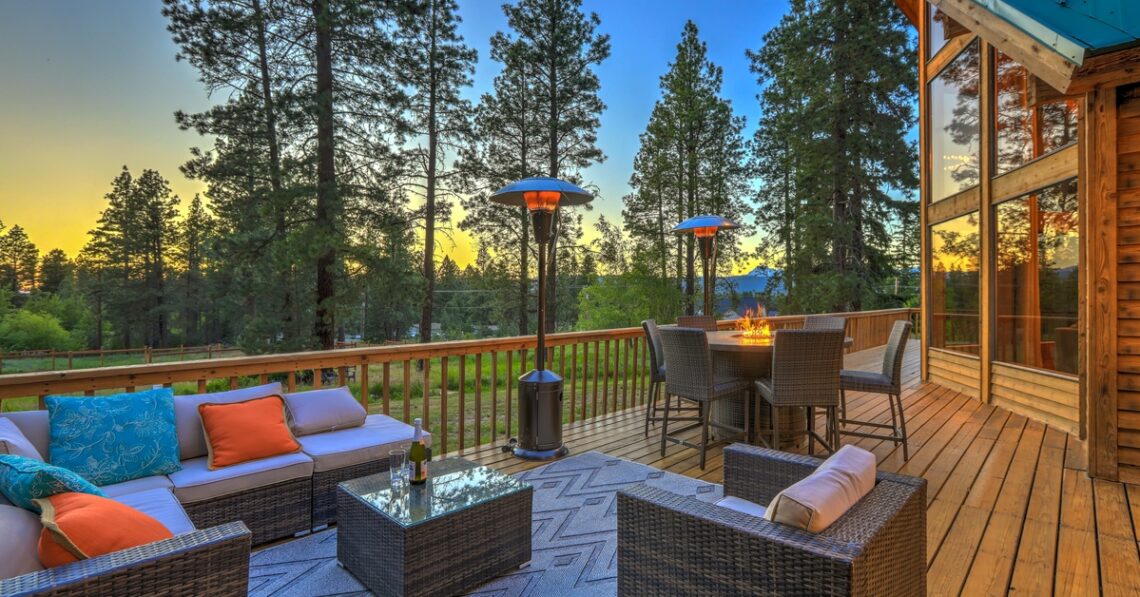 A cabin deck features a couch, several chairs, and a table with a fire. It sits next to a forest with tall trees.
