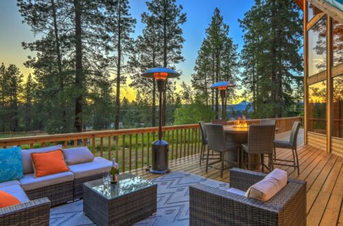 A cabin deck features a couch, several chairs, and a table with a fire. It sits next to a forest with tall trees.