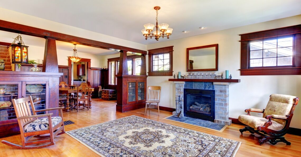 Warm living room with hardwood floors, stone fireplace, and dark wood mantel; a patterned area rug anchors the seating area beneath a chandelier and large windows with wooden trim.