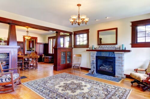 Warm living room with hardwood floors, stone fireplace, and dark wood mantel; a patterned area rug anchors the seating area beneath a chandelier and large windows with wooden trim.