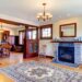 Warm living room with hardwood floors, stone fireplace, and dark wood mantel; a patterned area rug anchors the seating area beneath a chandelier and large windows with wooden trim.