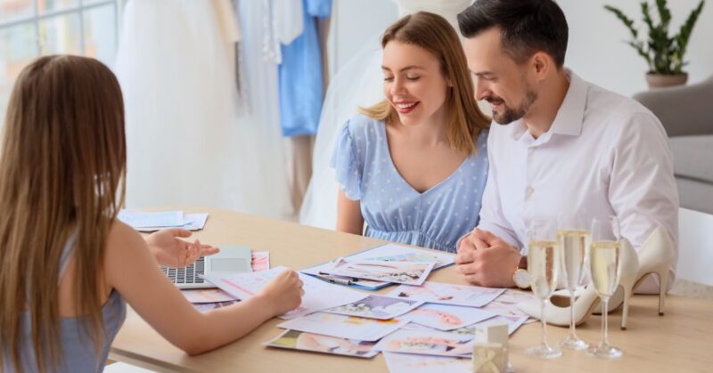 A couple sitting at a table with a woman while looking at planning sheets. There are champagne glasses next to them.
