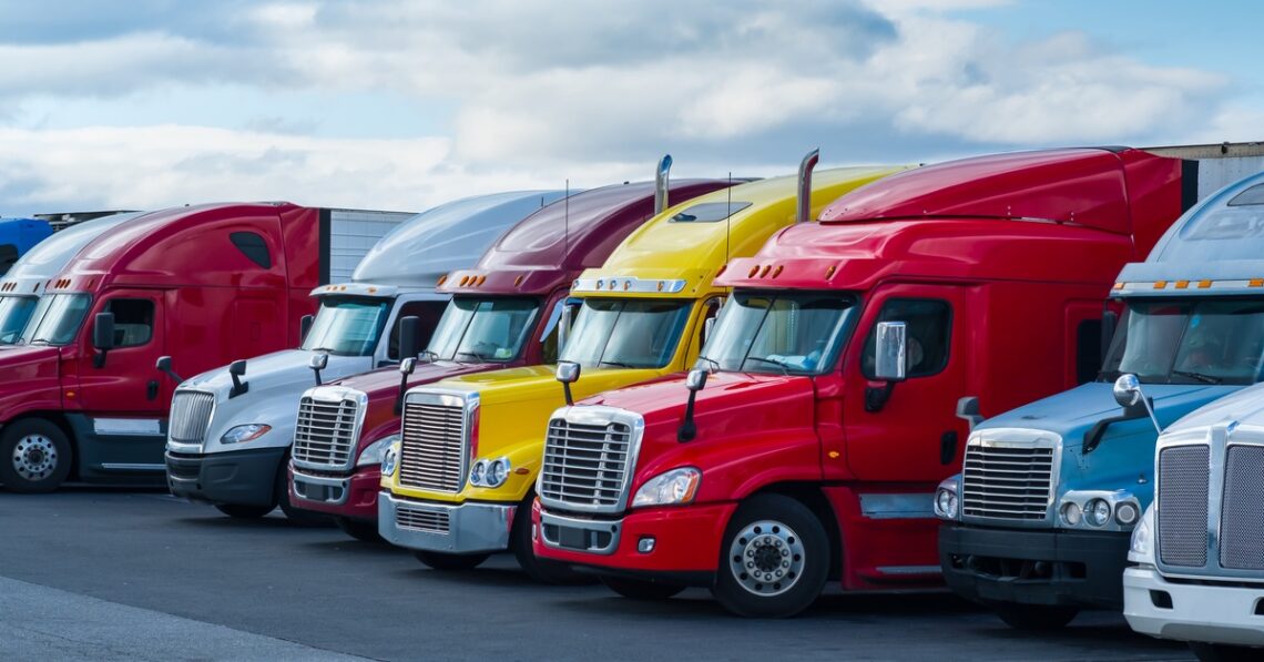 A row of colorful semi-trucks and trailers is parked on the pavement in a transportation lot under a bright sky.