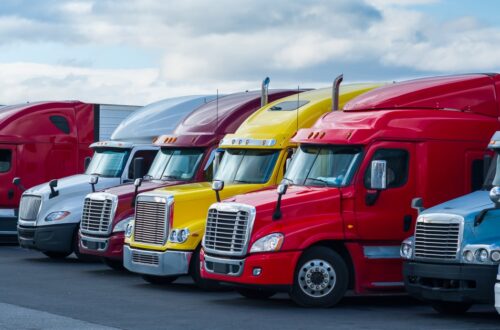 A row of colorful semi-trucks and trailers is parked on the pavement in a transportation lot under a bright sky.
