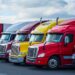 A row of colorful semi-trucks and trailers is parked on the pavement in a transportation lot under a bright sky.