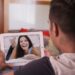 A man holds a tablet showing a smiling woman on a video call while sitting in a living room with pillows nearby.