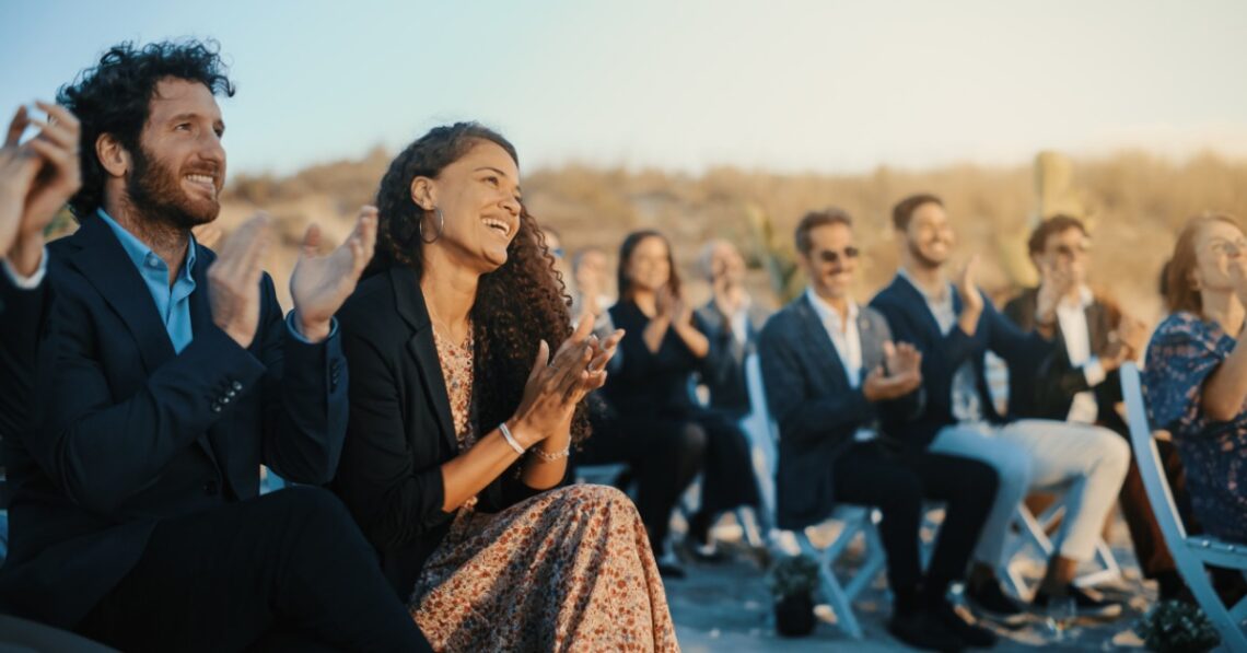Many well-dressed guests sit on white chairs outdoors, smiling and applauding during a sunlit wedding ceremony.