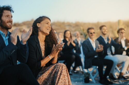 Many well-dressed guests sit on white chairs outdoors, smiling and applauding during a sunlit wedding ceremony.