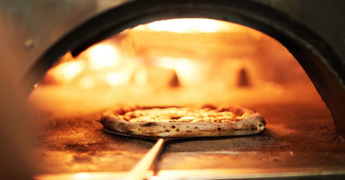 A chef uses a pan to retrieve a crispy pizza from a commercial-grade oven with a wood fire stoking inside.