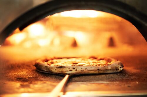 A chef uses a pan to retrieve a crispy pizza from a commercial-grade oven with a wood fire stoking inside.