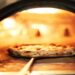 A chef uses a pan to retrieve a crispy pizza from a commercial-grade oven with a wood fire stoking inside.