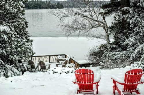 Two wooden Adirondack chairs sitting in snow near a frozen lake with bright sky, trees, and distant hills.
