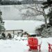 Two wooden Adirondack chairs sitting in snow near a frozen lake with bright sky, trees, and distant hills.