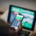 Mature man using mobile app for live betting and online gambling and watching a sports match Informed wagers