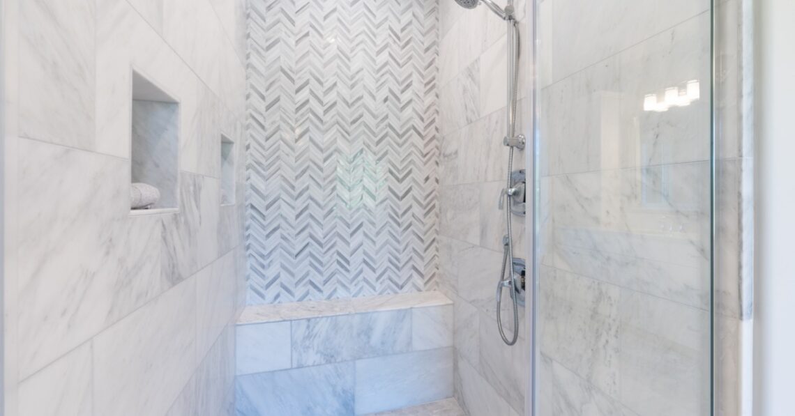 A walk-in shower covered in white and grey tile with a patterned back wall and a small concrete bench in the back.