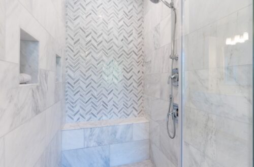 A walk-in shower covered in white and grey tile with a patterned back wall and a small concrete bench in the back.
