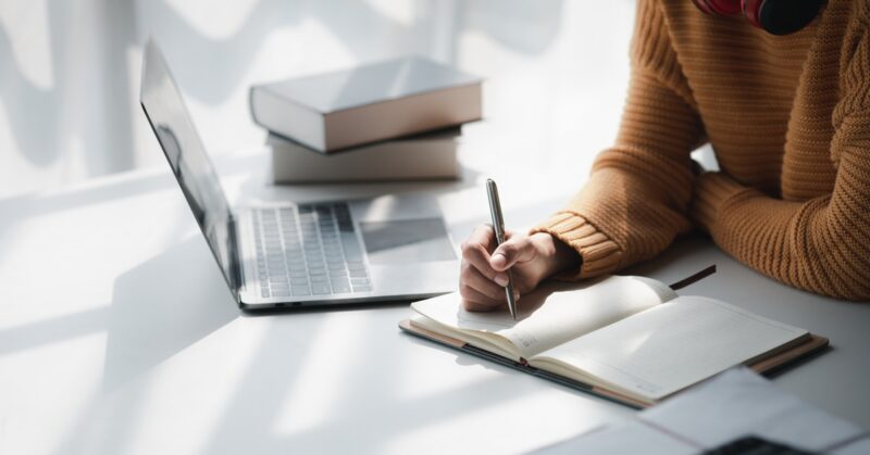 Person takes notes in a notebook while working beside an open laptop, books, and headphones at a bright desk.Homework Space