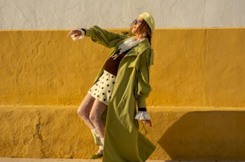 A woman wears a green coat with a polka-dot skirt and matching hat while posing against a yellow and white wall.