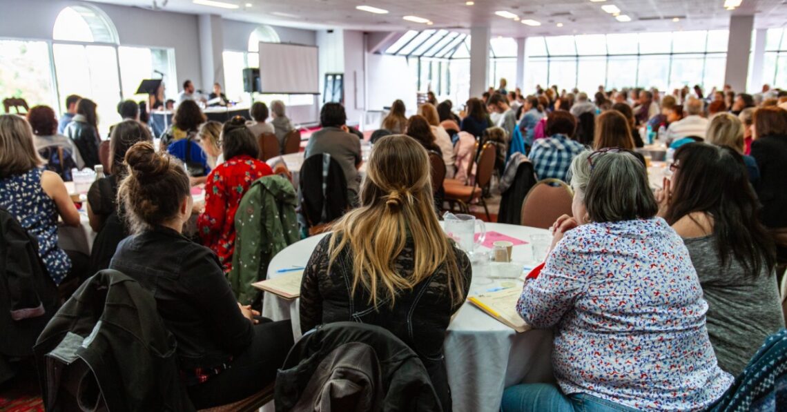A crowd of people sitting at round tables in front of a stage full of presenters at a corporate conference.