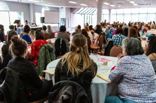 A crowd of people sitting at round tables in front of a stage full of presenters at a corporate conference.